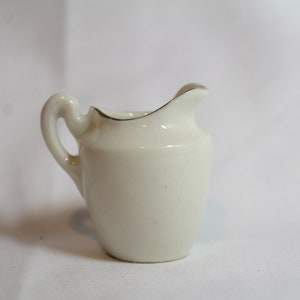 May include: A small, off-white ceramic creamer with a curved handle and a slightly flared spout. The rim is accented with a thin gold band. The creamer is designed for serving cream or milk.