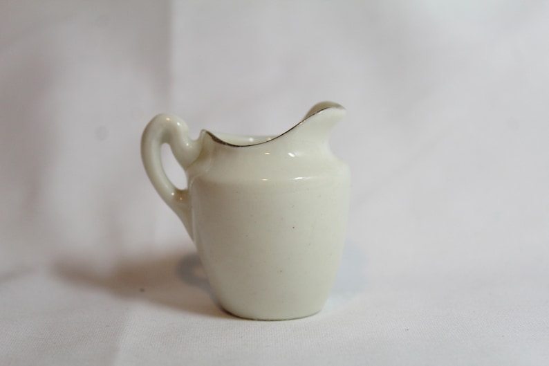 May include: A small, off-white ceramic creamer with a curved handle and a slightly flared spout. The rim is accented with a thin gold band. The creamer is designed for serving cream or milk.
