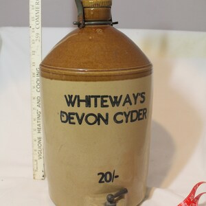 Puede incluir: Una jarra de cerámica marrón con una asa de metal y un grifo. La jarra tiene una etiqueta que dice "WHITEWAY'S DEVON CYDER 20/-."