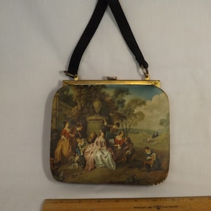 Op de afbeelding: Een vintage handtas met een zwarte band en een gouden sluiting. De voorkant van de tas is bedekt met een bedrukte stof die een scène van mensen laat zien die samenkomen in een tuin.