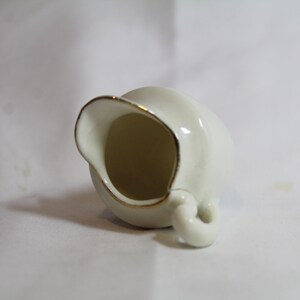 May include: A small, off-white ceramic creamer with a gold rim and handle. The creamer is tilted, showcasing its interior. The design is simple and elegant, suitable for serving cream or milk. The creamer is likely part of a tea or coffee set.