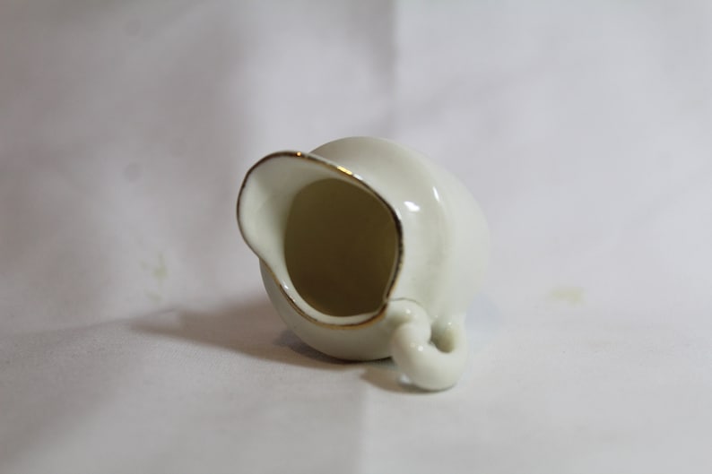 May include: A small, off-white ceramic creamer with a gold rim and handle. The creamer is tilted, showcasing its interior. The design is simple and elegant, suitable for serving cream or milk. The creamer is likely part of a tea or coffee set.