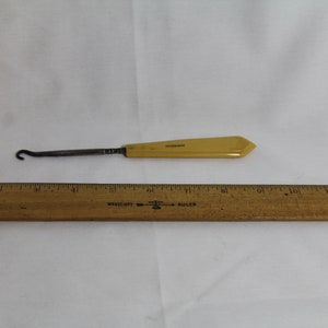 Peut inclure: Un chausse-pied en plastique jaune avec un crochet en métal. Le chausse-pied mesure environ 15 cm de long et est présenté à côté d'une règle en bois.