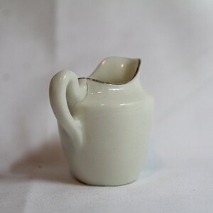 May include: A small, off-white ceramic creamer with a curved handle and a spout. The rim is accented with a thin gold band. The creamer is a classic design, suitable for serving cream or milk. It is a traditional piece for a tea or coffee service.