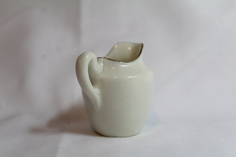 May include: A small, off-white ceramic creamer with a curved handle and a spout. The rim is accented with a thin gold band. The creamer is a classic design, suitable for serving cream or milk. It is a traditional piece for a tea or coffee service.
