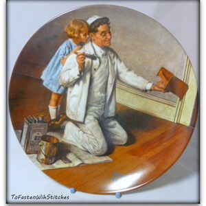 May include: A vintage ceramic plate depicting a painting of a man and a young girl. The man is kneeling and holding a paintbrush, while the girl stands beside him. The plate has a brown wooden background and a white border.