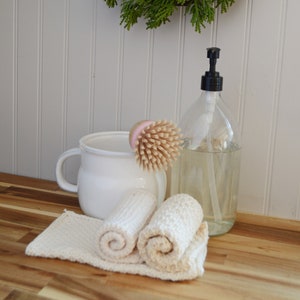 May include: Three white crocheted dishcloths, a white ceramic jug with a handle, a wooden brush with pink bristles, and a clear glass bottle with a black pump dispenser. The items are arranged on a wooden worktop. The background is a white panelled wall with a green wreath hanging on it. The text "CROCHET PATTERN" is visible above the wreath.
