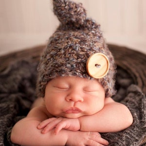 May include: A newborn baby wearing a brown and blue crocheted hat with a wooden button. The baby is sleeping with their arms crossed and their head resting on their arms.