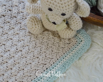 May include: A crocheted baby blanket in a beige color with a light blue border. The blanket is made with a textured stitch pattern and is lying on a white fluffy surface. A crocheted teddy bear is sitting on top of the blanket.