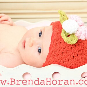 May include: A baby wearing a red crocheted hat with a pink and green flower on top. The hat is in the shape of a strawberry.
