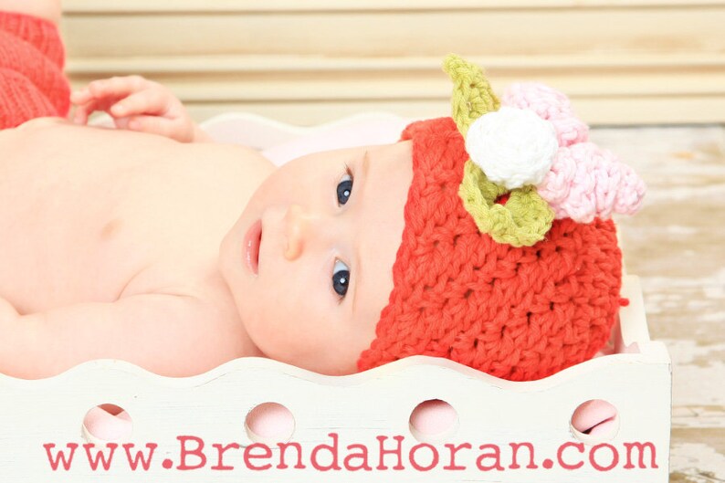 May include: A baby wearing a red crocheted hat with a pink and green flower on top. The hat is in the shape of a strawberry.