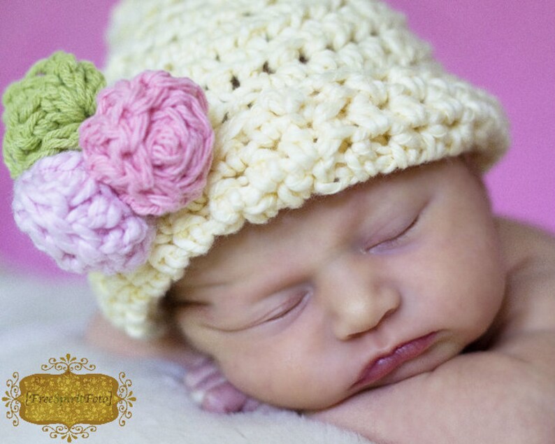 May include: A sleeping baby wearing a crocheted hat with pink and green flower embellishments.