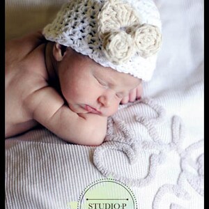 May include: A sleeping baby wearing a white crocheted hat with a beige crocheted flower accent. The baby is lying on a white blanket with an embroidered design.