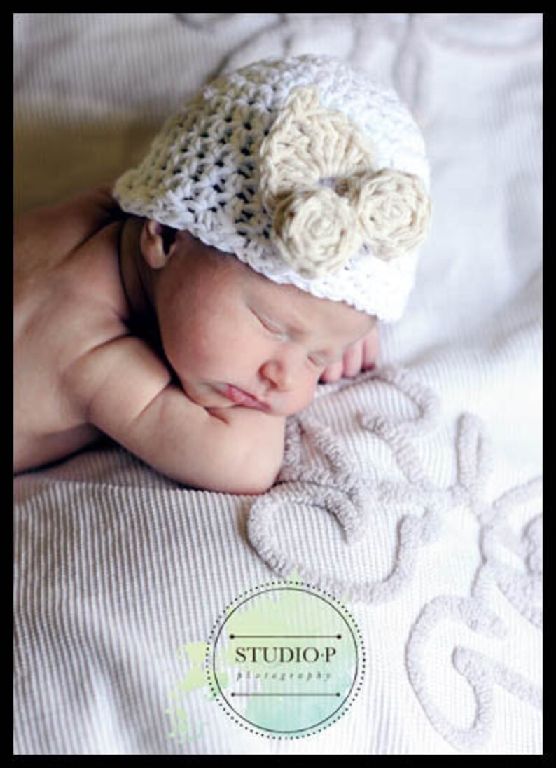 May include: A sleeping baby wearing a white crocheted hat with a beige crocheted flower accent. The baby is lying on a white blanket with an embroidered design.