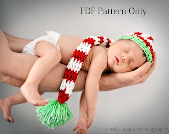 May include: A sleeping baby wearing a red, white and green crocheted hat and scarf. The baby is being held by an adult's arm.