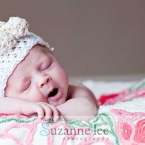 May include: A baby wearing a white crocheted hat with a flower design sleeps on a white and pink blanket with floral embroidery.