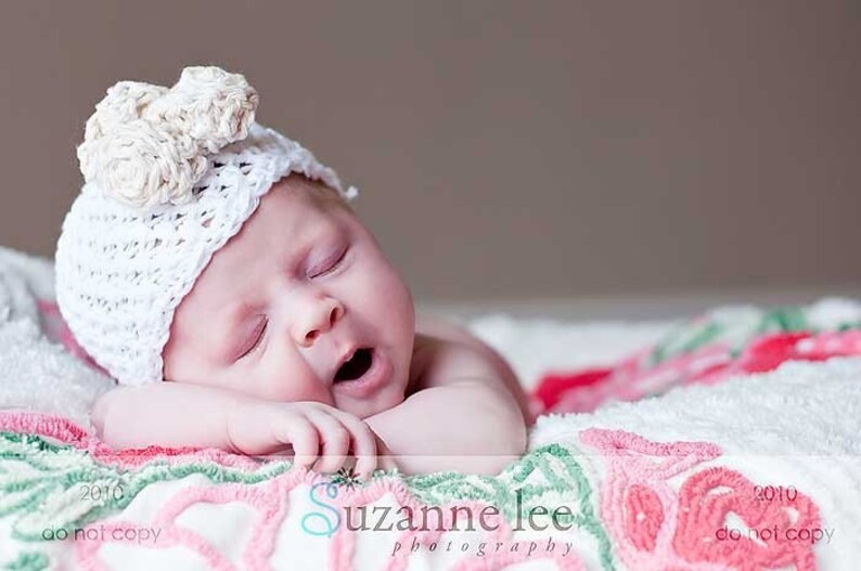 May include: A baby wearing a white crocheted hat with a flower design sleeps on a white and pink blanket with floral embroidery.