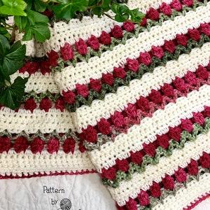 May include: A white, green, and pink crocheted blanket with a striped pattern. The blanket is made with a textured stitch that creates a raised effect. The text "Pattern by Babies by Hand Props and Patterns" is visible in the bottom left corner of the image.
