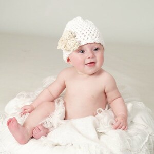 May include: A baby wearing a white crocheted hat with a beige flower accent sits on a white, lacy blanket.
