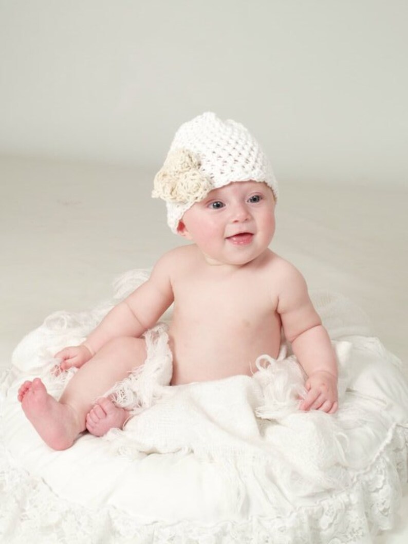 May include: A baby wearing a white crocheted hat with a beige flower accent sits on a white, lacy blanket.