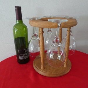May include: A wooden wine glass holder with six glasses hanging from it. A bottle of wine is sitting on a red tablecloth next to the holder.