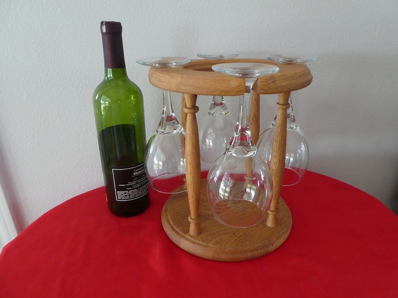 May include: A wooden wine glass holder with six glasses hanging from it. A bottle of wine is sitting on a red tablecloth next to the holder.
