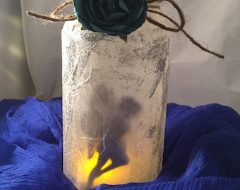 May include: A decorative glass jar with a fairy silhouette illuminated by a warm light. The jar is embellished with silver glitter and a teal fabric flower. A twine bow adorns the top, creating a whimsical, handcrafted aesthetic.