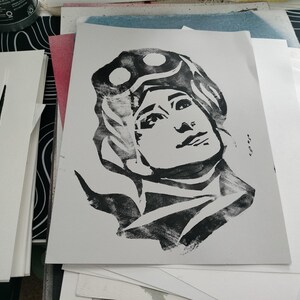 May include: Black and white print of a person's face with a helmet or goggles on their head. The print is on a white background.
