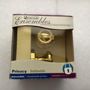 May include: A gold-toned privacy locking door knob set by Amerock Ensembles in its original packaging. The box features the text Amerock Ensembles, Privacy, Intimité, and Bedroom/Bath, with a blue icon of a padlock. The knobs have a polished finish.