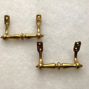 May include: Two polished brass drawer pulls featuring a horizontal bar with a central decorative bead and pointed ends. Each pull has two vertical mounting brackets, each containing two circular screw holes for attachment to furniture surfaces.