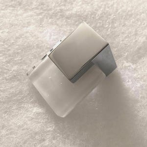 May include: A modern, geometric ring featuring a clear, rectangular base topped with a silver-toned, rectangular accent. The ring has a minimalist design and a polished finish, suitable for everyday wear.