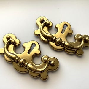 May include: Two antique brass drawer pulls with a decorative, curved design. Each pull features a keyhole and a curved handle with a series of small, round details. The pulls are a warm, golden color.
