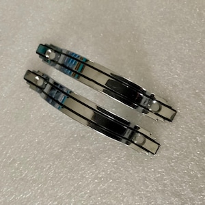 May include: Two shiny, silver metal hair barrettes with a curved design. Each barrette has a black stripe down the center and small, decorative accents. The barrettes are displayed on a textured, white surface.