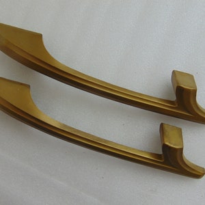 May include: Two matching gold-toned cabinet pulls with a curved, elongated design. Each handle features a smooth, matte finish and a distinct, tapered shape that extends from a rectangular mounting point, creating a sleek and uniform appearance.