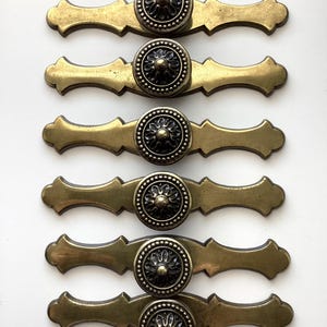 May include: Six antique brass drawer pulls with a decorative floral design. Each pull features a central circular knob with a detailed pattern, surrounded by a series of small dots. The pulls are approximately 6 inches long, with a curved, ornate shape.