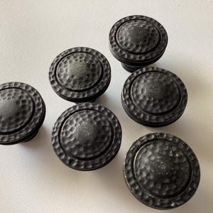 May include: Six dark gray cabinet knobs with a hammered texture. The knobs are round with a slightly raised center and a textured rim. These hardware pieces are suitable for furniture or cabinet upgrades.