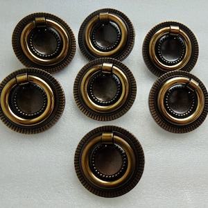 May include: Seven antique brass cabinet pulls with ring handles. Each pull has a circular design with a ridged outer edge and a beaded inner ring. The pulls are arranged on a white surface.