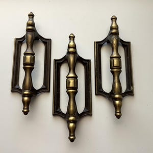 May include: Three vintage bronze-toned door pulls. Each pull features a central, ornate handle within a rectangular frame. The metal has a warm, aged appearance, suggesting a classic or antique style. The pulls are likely for furniture or doors.