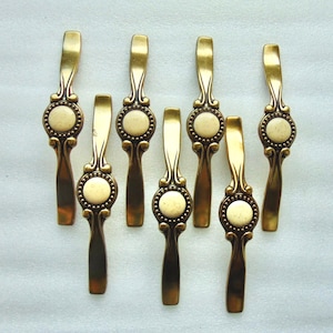 May include: A set of seven vertical cabinet pulls featuring a metallic antique brass finish. Each pull has a central circular medallion with a speckled cream-colored inlay, surrounded by decorative beaded detailing and curved metal extensions.
