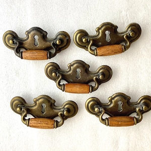 May include: Five antique brass drawer pulls with keyhole details and wooden knobs. The pulls have a decorative, ornate design.