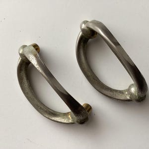 May include: Two antique silver-toned drawer pulls with a D-shaped design. Each pull has a rounded handle and is attached with small brass-colored screws. The pulls are likely for a cabinet or drawer.