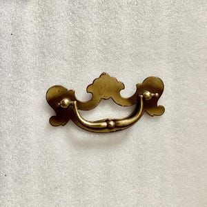 May include: A gold-toned metal drawer pull with a decorative, ornate design. The pull has a curved handle and two small knobs on the sides.