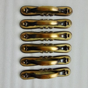 May include: Six antique brass cabinet pulls. Each pull has a curved handle with decorative detailing and is mounted on a rectangular backplate. The pulls have a vintage, aged appearance, with a mix of gold and dark brown tones.
