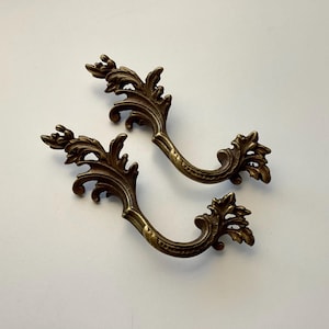 May include: Two ornate, antique-style bronze drawer pulls. Each pull features a curved handle with intricate leaf and scroll detailing. The bronze finish has a warm, aged appearance, suggesting vintage or classic furniture hardware.
