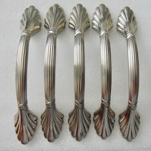 May include: Five silver-toned cabinet handles. Each handle features a curved, ribbed center section and decorative shell-shaped ends. The handles are made of metal and are designed for furniture or cabinetry.