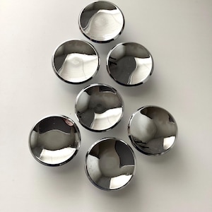 May include: Seven shiny, chrome-finished cabinet knobs arranged on a white surface. Each knob is round with a slightly concave center, reflecting the surrounding environment. The knobs are evenly spaced, creating a visually appealing pattern.