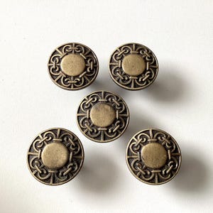 May include: Five antique brass cabinet knobs with a circular design. Each knob features a central circle surrounded by an ornate, repeating pattern. The knobs are a warm, aged brass color, and are set against a white background.