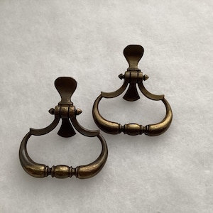 May include: Two matching brass drawer pulls with a dark, aged patina. Each pull features a vertical mounting plate with a flared top and a hinged, curved handle that swings downward, showcasing a central decorative bead detail.