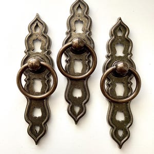 May include: Three antique bronze-colored drawer pulls. Each pull features an ornate, elongated backplate with a decorative cutout design and a round ring handle. The hardware is likely for cabinets or furniture, adding a vintage aesthetic.