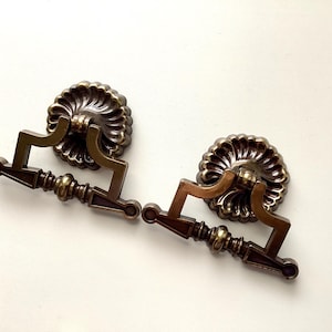 May include: Two antique bronze drawer pulls. Each pull features a decorative, fluted backplate and a handle with a detailed, geometric design. The pulls are a warm, metallic color, suggesting a vintage aesthetic.